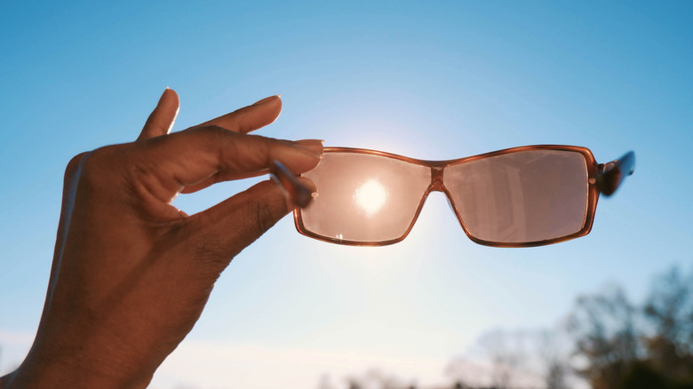 A woman's hand holds sunglasses up against the sky with the sun visible through lens