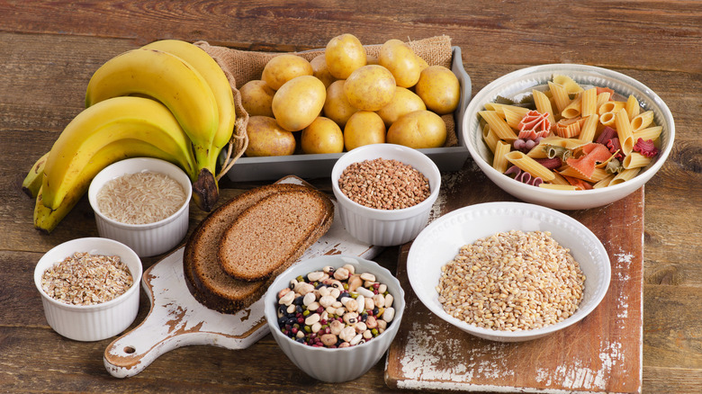 Assorted high carbohydrate foods on a table, including bananas, potatoes, pasta, rice, and bread