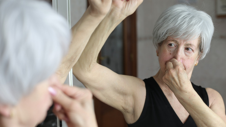 Senior woman standing in the mirror with one arm raised and the other hand holding her nose