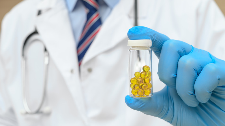 A doctor in a white lab coat and tie holds a vial of medicine in his blue-gloved hand