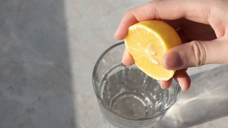 A close-up of a hand squeezing half of a lemon over a glass of water.