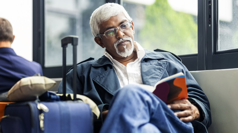 Man with white hair and beard reading a book while seated waiting in an airport