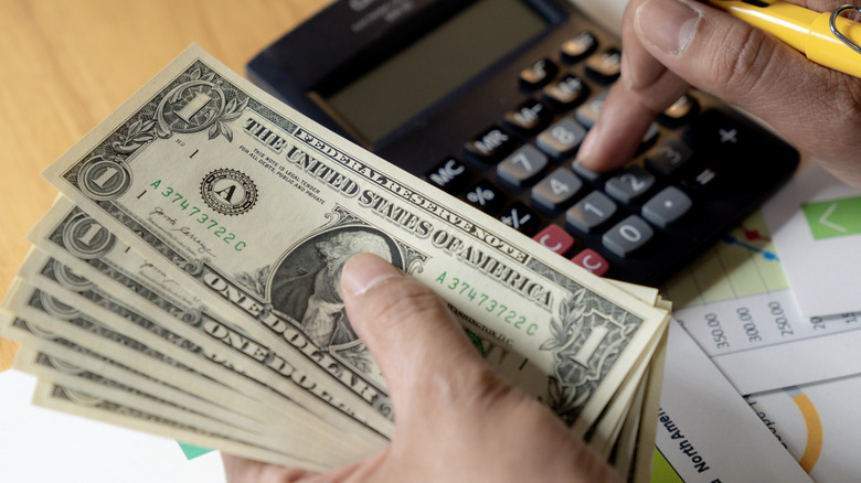 A person sits at a table, holding money in one hand and using a calculator with the other