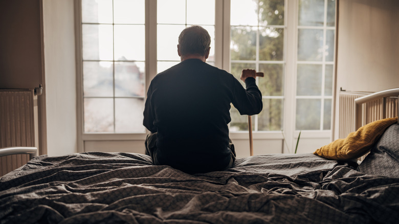 A lonely man sits on a bed facing a window, holding a cane in his hand