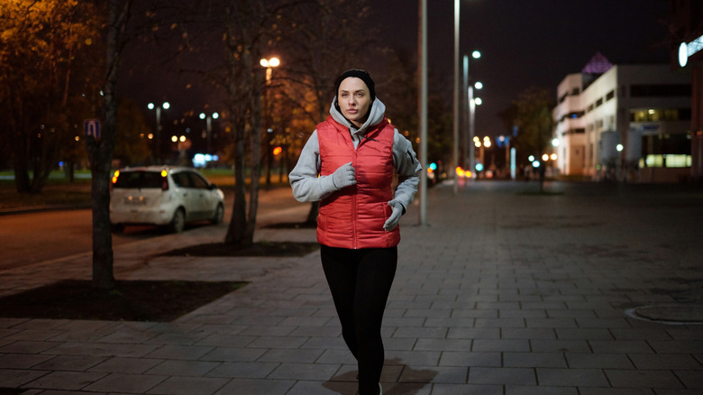 Woman in gray hoodie and red vest running on a sidewalk in the evening
