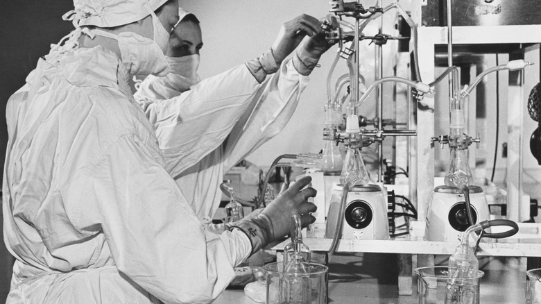 A black and white image of two scientists working with lab equipment while wearing safety gear.
