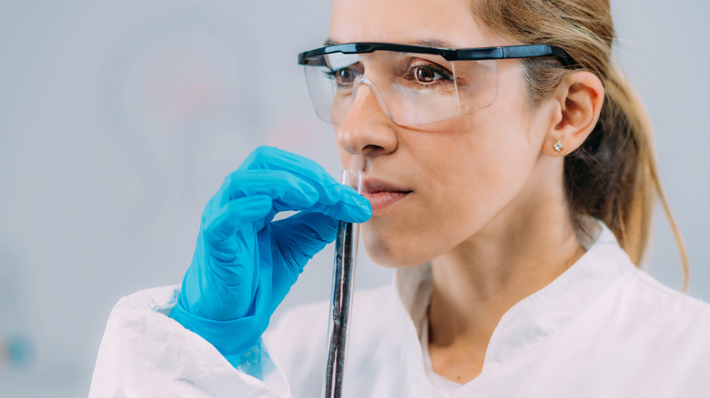 Woman in lab coat with gloves and safety glasses smelling a vial