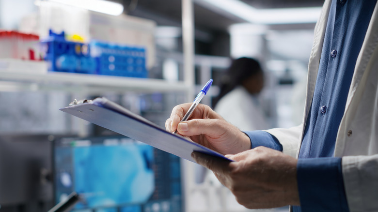 A man in a lab coat stands in front of a blurred lab bench while taking notes on a clipboard.