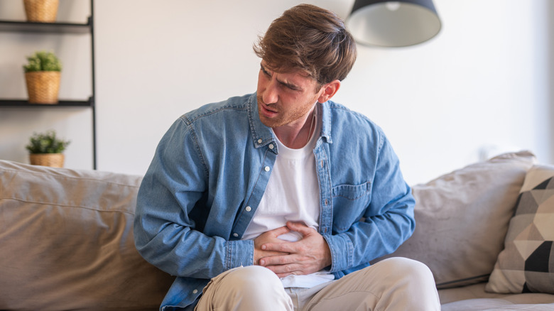 A man in a blue shirt sitting on a sofa and holding his abdomen in pain