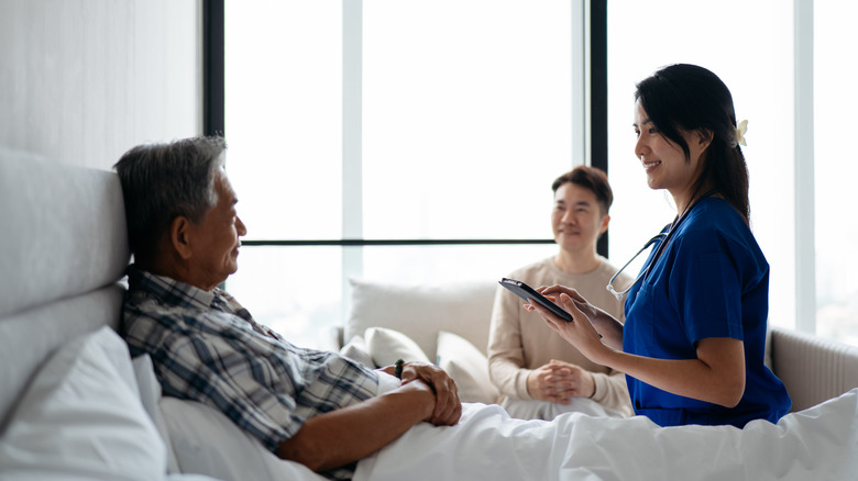 A healthcare worker sits on a bed, speaking to an elderly man who is sitting up in the bed.