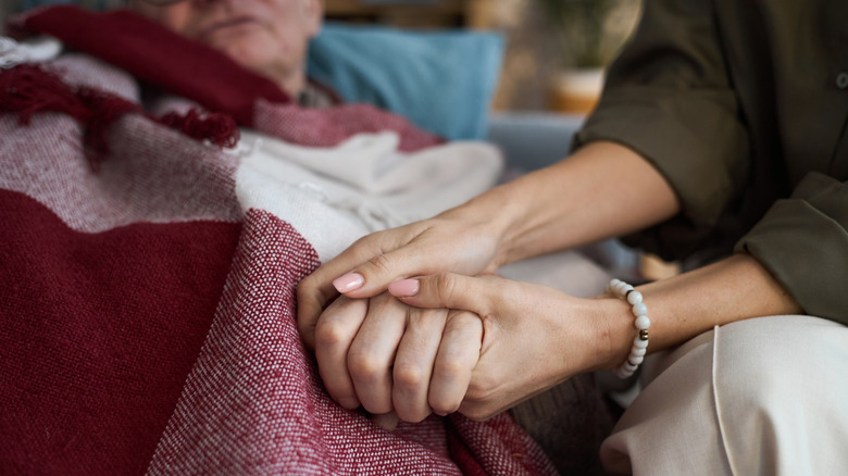 A close-up of a woman holding a man's hand while he lays in a bed.