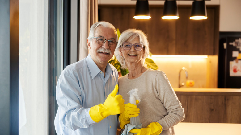 Senior couple with thumbs up and rubber gloves and cleaning supplies