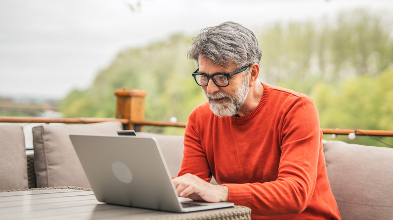 Older white grey-bearded man with glasses sitting in patio with laptop