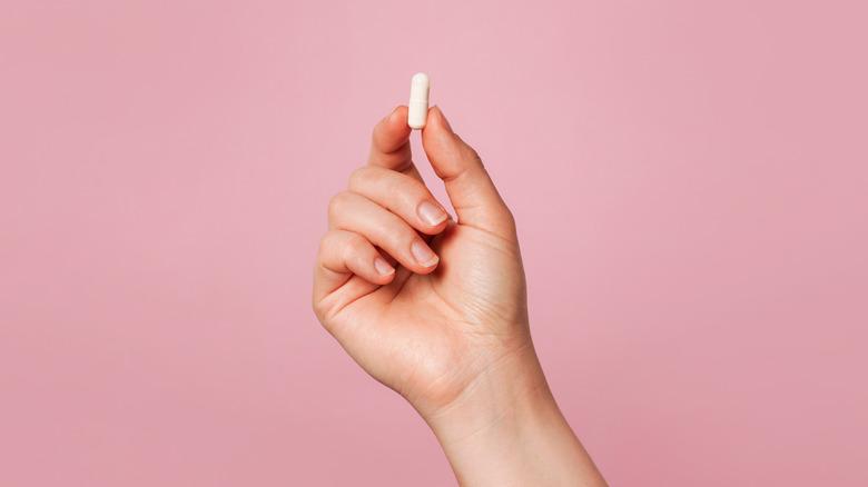 A hand holding a white pill in front of a pink background.