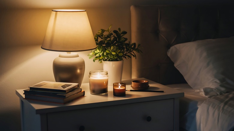 A bedside table with books, a plant, an illuminated lamp, and two lit candles.