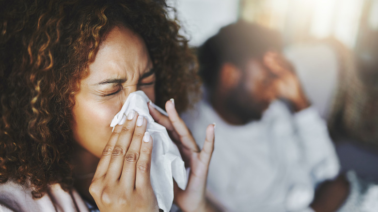 Woman blowing her nose with man in background with hand on head