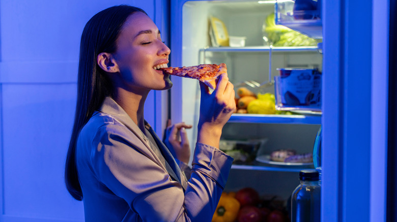 Woman eating a slice of pizza in front of open fridge