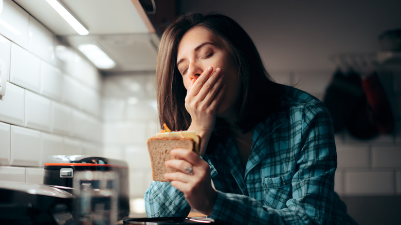 Woman holding a sandwich while yawning in kitchen