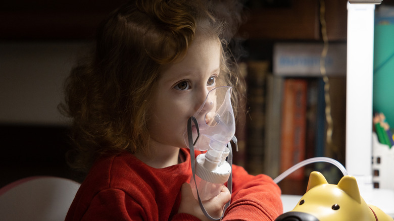 A little girl holds a nebulizer to her face while sitting beside a toy dog.
