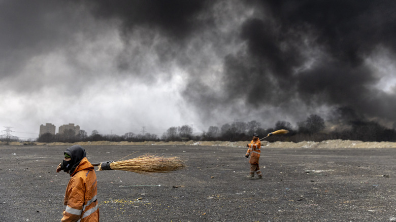 Billowing black smoke from airstrike on Iranian oil facilities as mask-clad workers cross an empty field