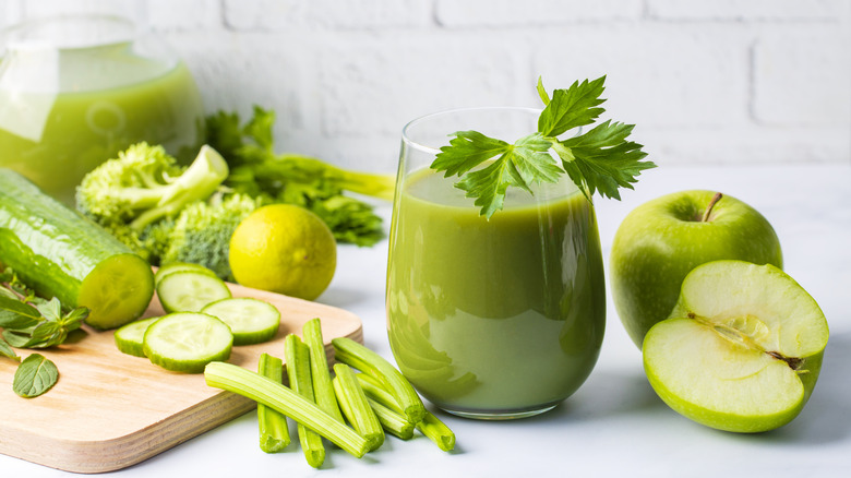 Green smoothie in a glass surrounded by broccoli, celery, cucumber, lime, herbs, and green apples