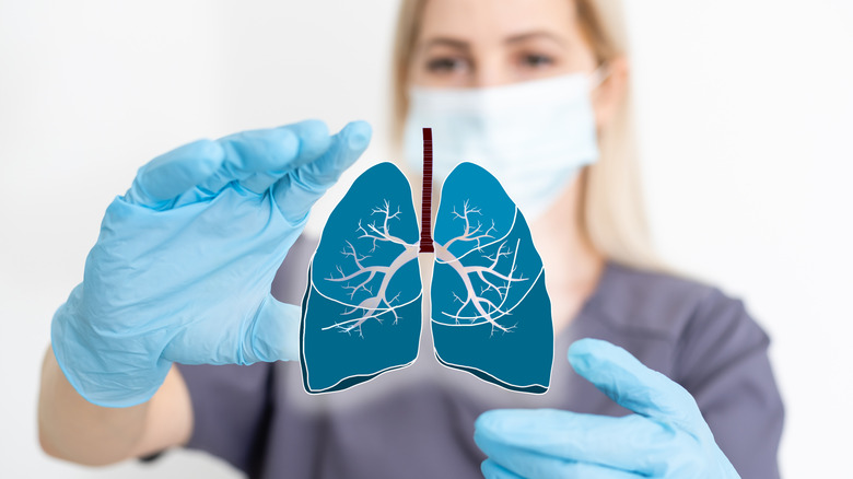Masked nurse in scrubs with gloved hands raised holding a cartoon set of lungs