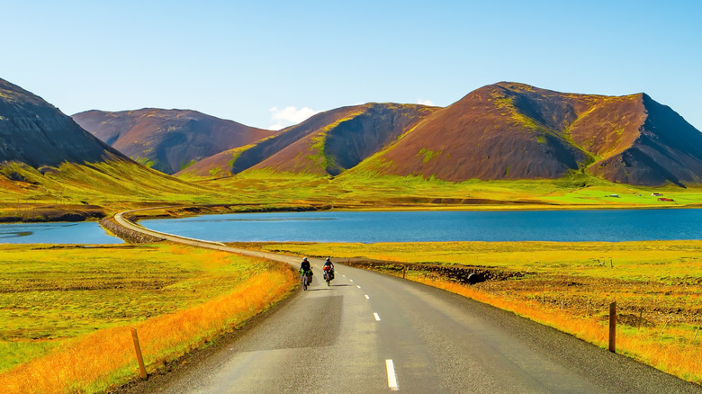 Iceland highway, two motorcyclists riding towards camera, treeless mountains and still lake in background