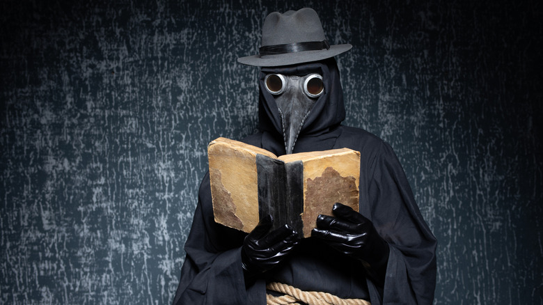 A plague doctor standing in front of a dark wall while reading a book.