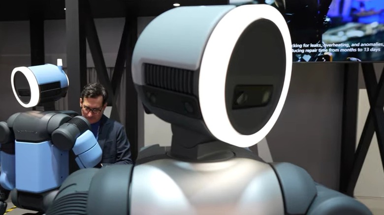 Close up image of circular head of Atlas robot, smooth plating, engineer working on second robot in background