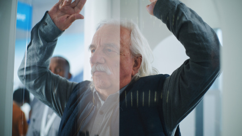 An elderly man holds his hands above his head while standing in an airport body scanner
