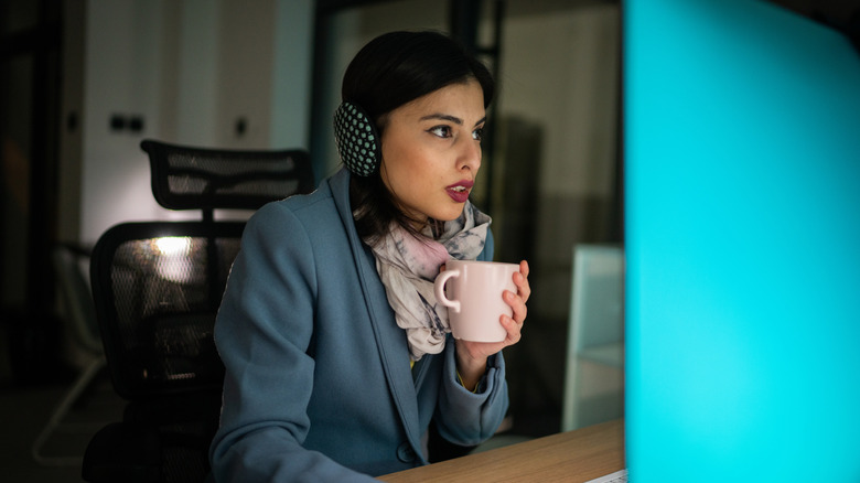 A woman at her desk with a scarf, ear muffs, and a cup of coffee