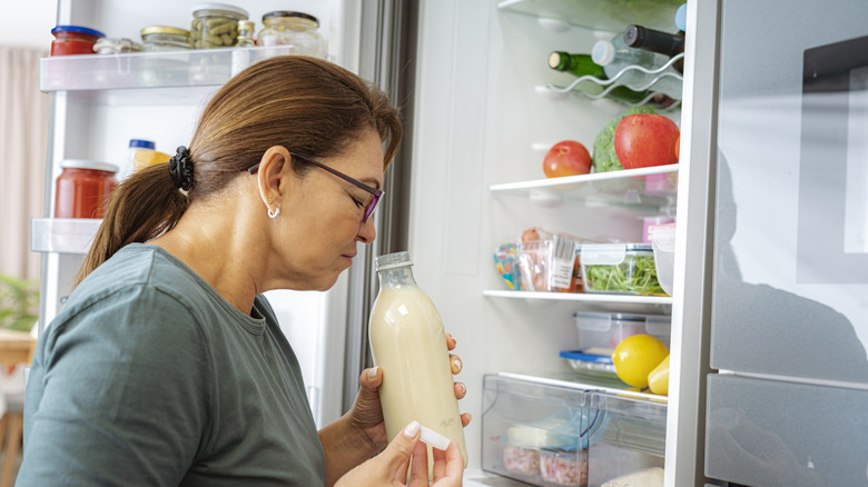 Woman standing in front of open refrigerator, smelling bottle of milk with a look of disgust on her face