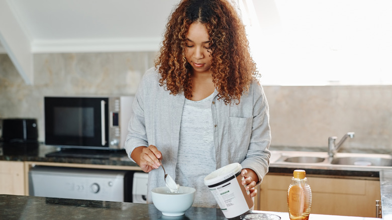 A woman making a yogurt bowl for breakfast, with a container of yogurt and bottle of honey on the counter
