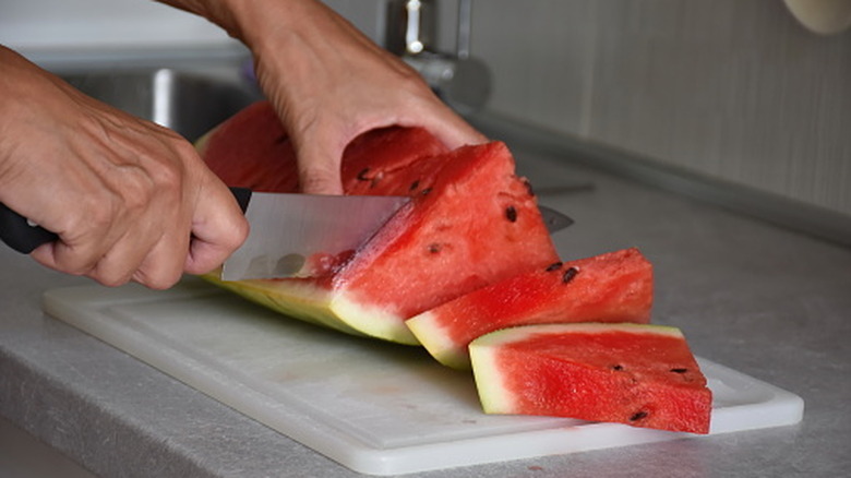 A person slicing watermelon on a white plastic cutting board with a knife