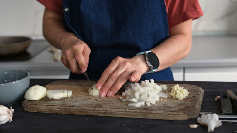 A woman wearing a blue apron cuts up an onion on a wooden cutting board