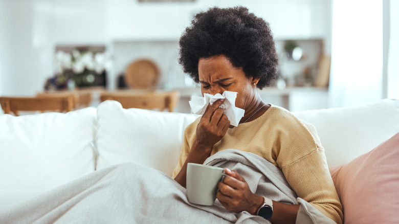 Woman blowing nose while sitting on sofa and holding mug
